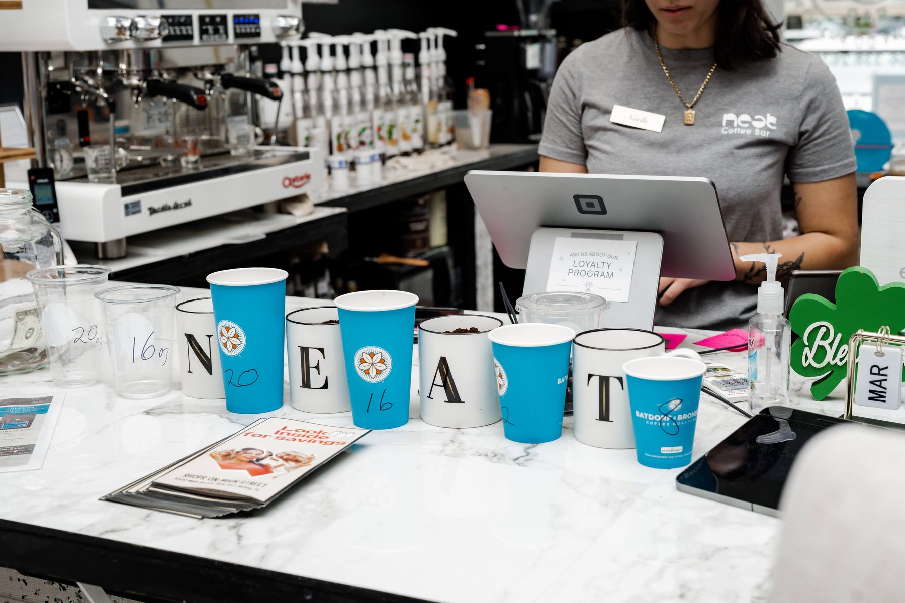 A barista at the counter with NEAT branded cups spelling out NEAT